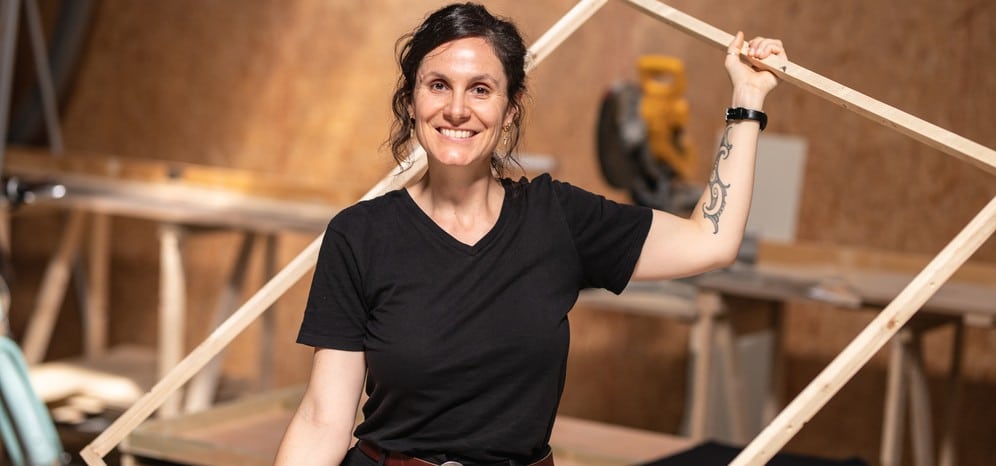 Une femme sourit dans un atelier de menuiserie, un cadre en bois brut posé sur son épaule.