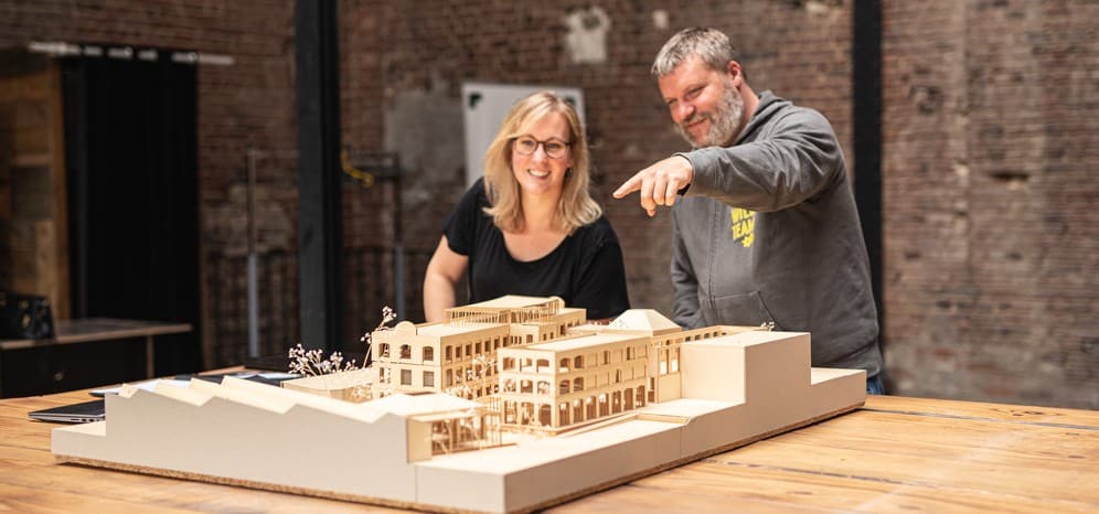 Deux personnes examinent la maquette d'un bâtiment posée sur une grande table, dans un ancien local industriel en brique.